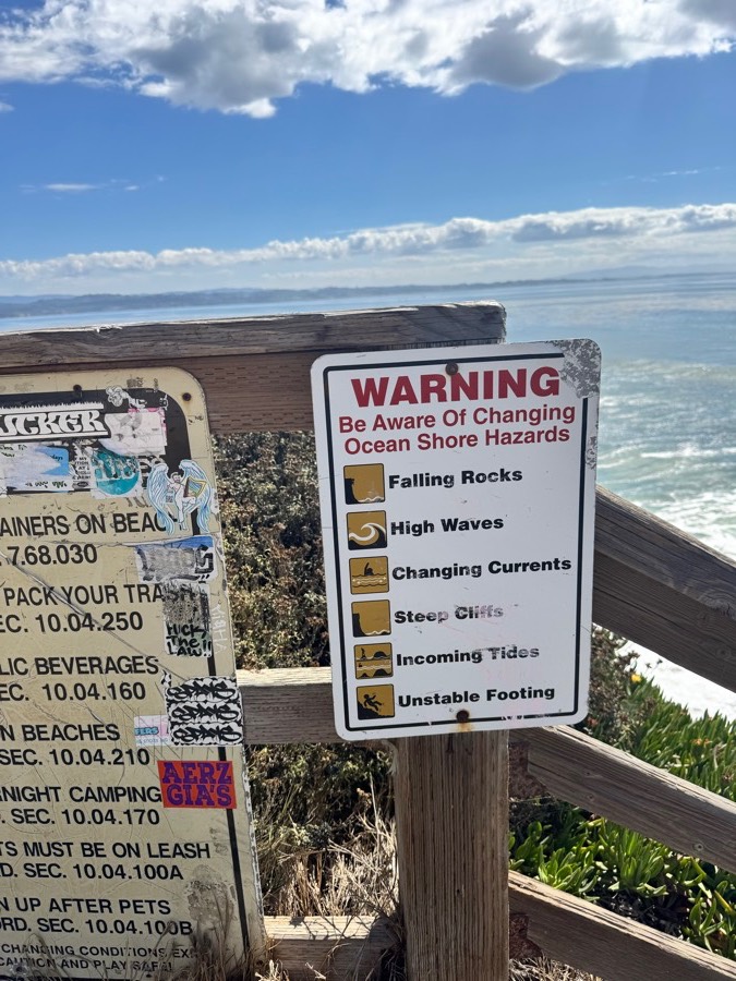 A white warning placard headed &ldquo;Be Aware Of Changing Ocean Shore Hazards&rdquo; listing, with pictograms, falling rocks, high waves, changing currents, steep cliffs, incoming tides and unstable footing. Beside it a heavily stickered county ordinance sign; behind, the ocean and a wooden rail.