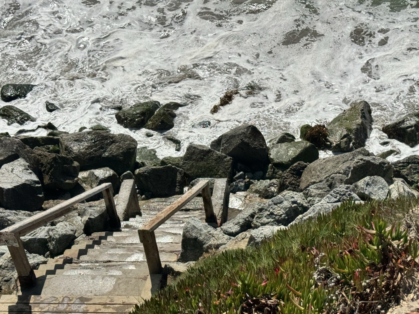 Concrete steps with weathered wooden handrails descend between large angular granite boulders into a frame filled with breaking whitewater. A dark algae band crosses the lower half of the boulders.